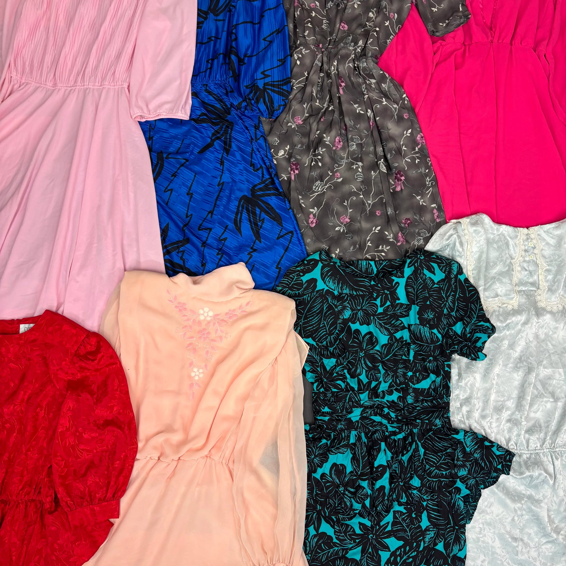 Assorted vintage 80s dresses in pink, blue, gray floral, hot pink, red, peach, teal floral, and white lace fabrics laid flat.