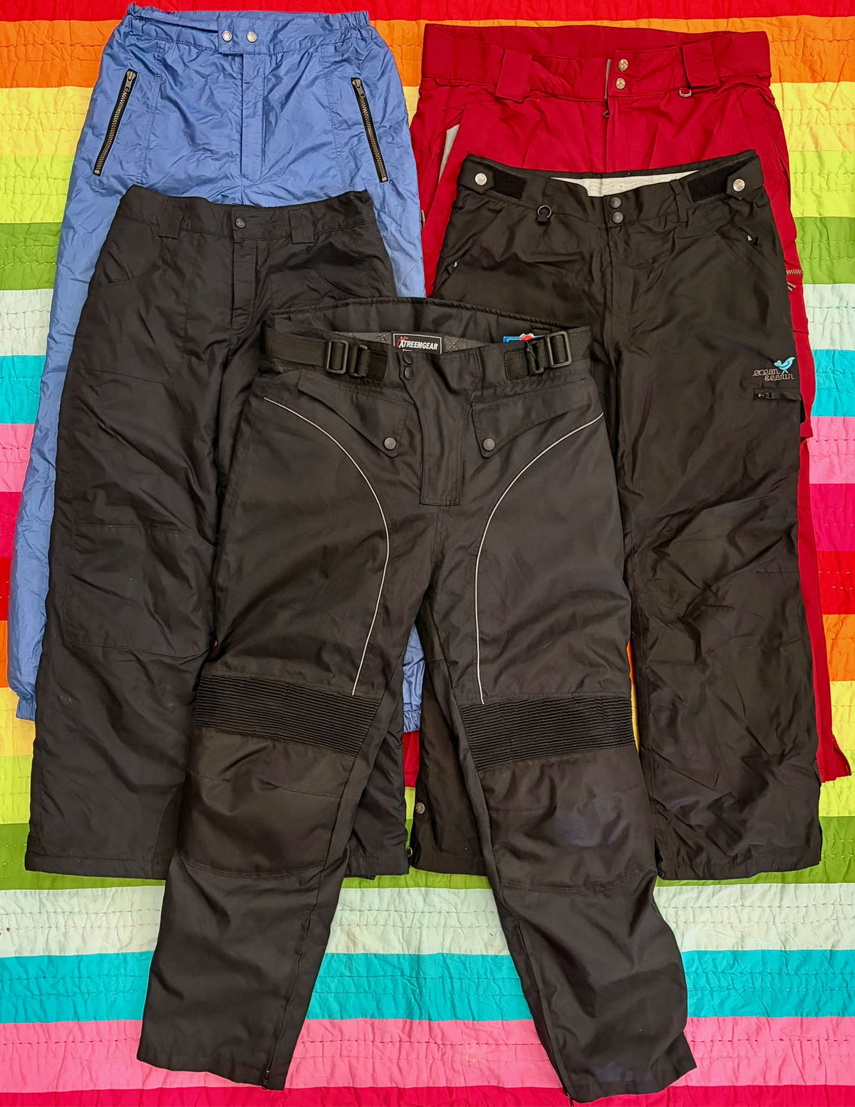 Three pairs of black and blue ski pants with reinforced knees, arranged on a colorful striped surface.