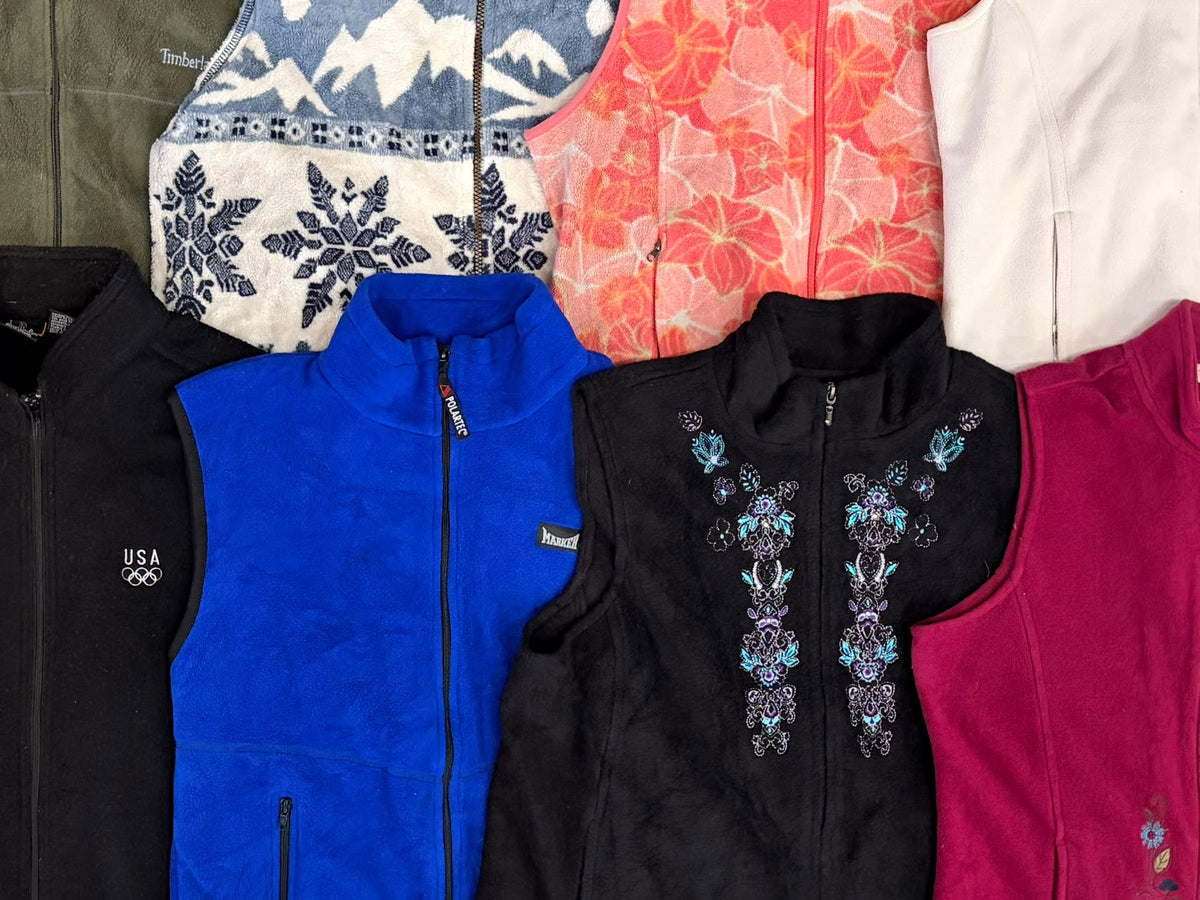 Assorted fleece gilets in black, bright blue, red, cream, and patterned designs, with zip fronts and embroidered details.